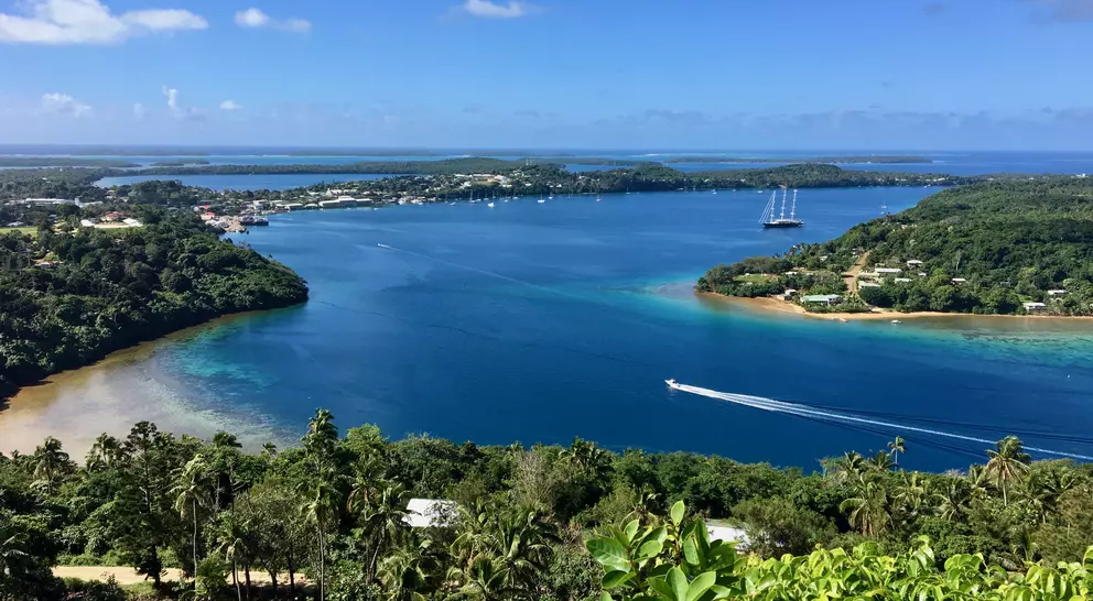A scenic view of a bay with clear blue water, surrounded by greenery and small boats, under a bright blue sky.