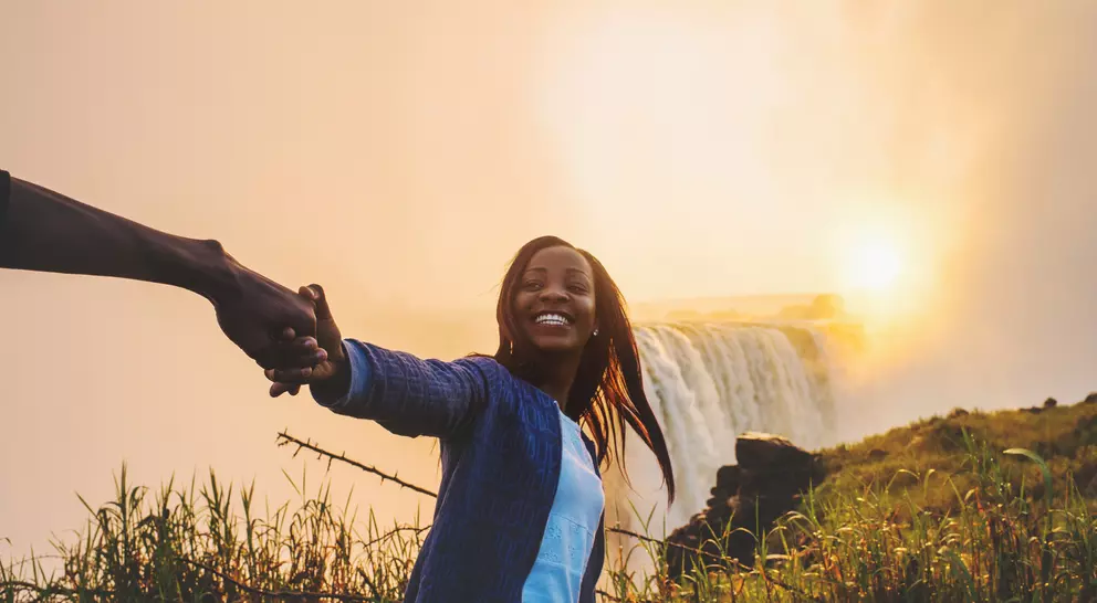 A woman joyfully holds hands with a person, standing near a waterfall at sunset amidst lush greenery.