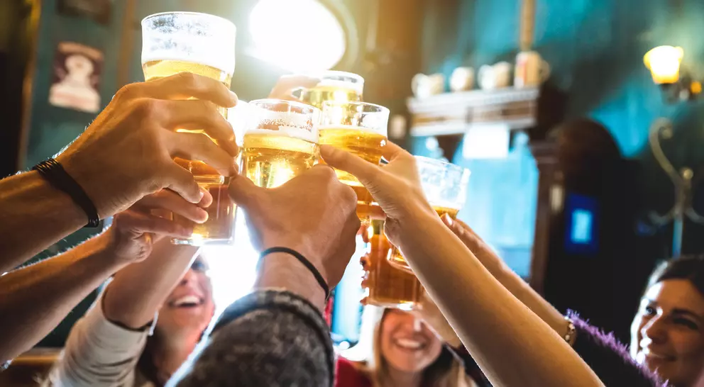 Group of people raising glasses of beer in a toast, smiling, inside a lively pub with colorful decor.