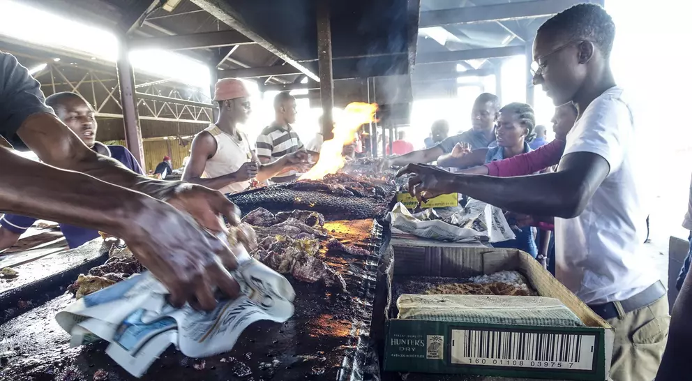A bustling market scene with people grilling food over open flames, surrounded by vendors and onlookers.