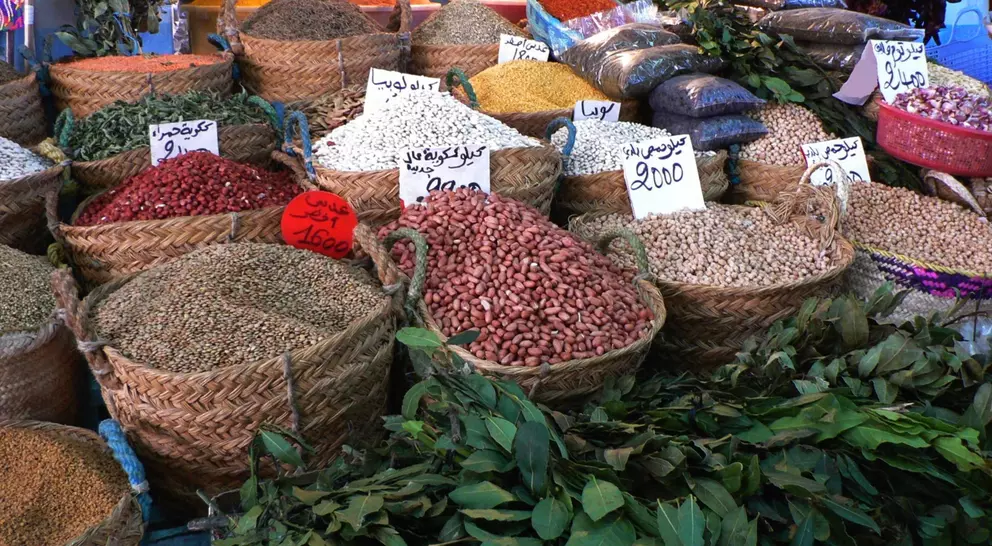 Baskets of colorful spices and legumes arranged in a market, with price tags visible in Arabic.