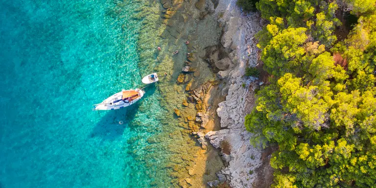 A small sailing boat is moored next to the shore which is covered with a lush pine forest in Hvar