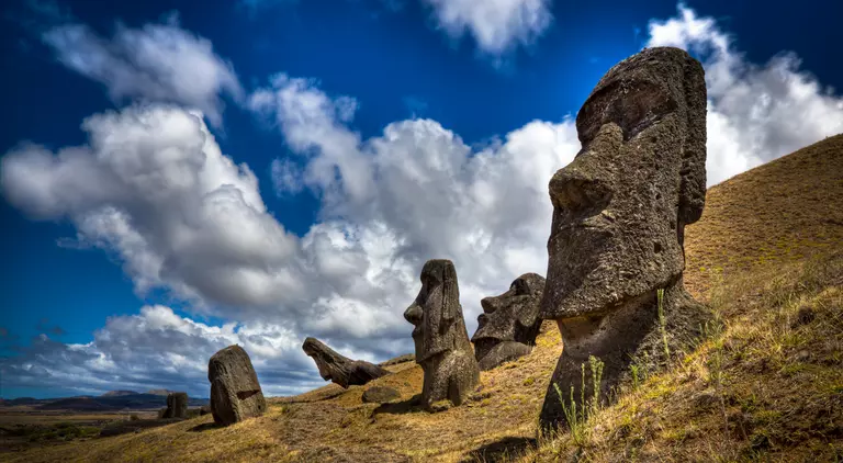 One Moais is in the foreground and four in the background. The sky is mostly sunny with clouds.