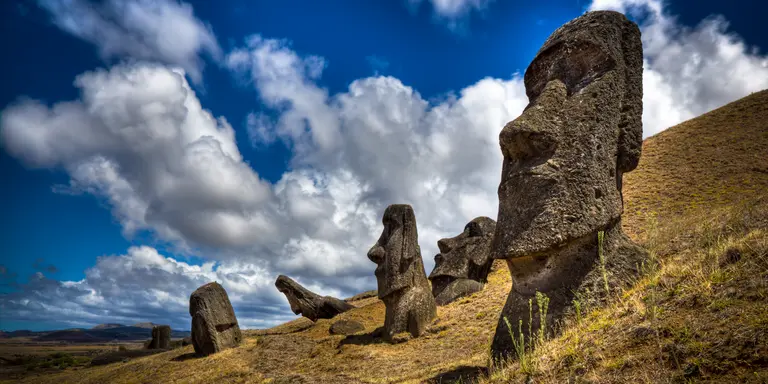 One Moais is in the foreground and four in the background. The sky is mostly sunny with clouds.