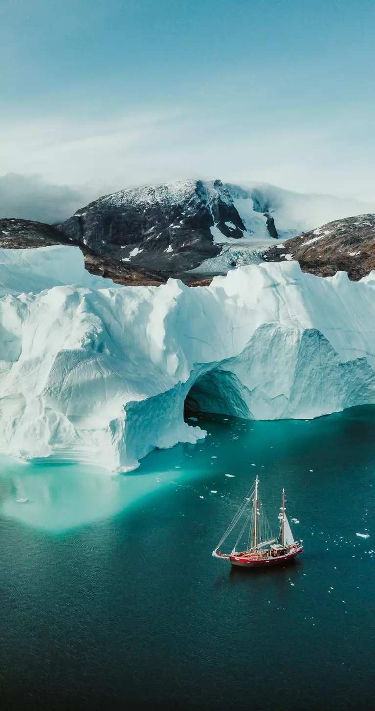 An aerial view of a sailboat passing alongside icebergs in the cold Arctic waters of Greenland.