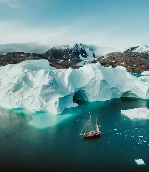 An aerial view of a sailboat passing alongside icebergs in the cold Arctic waters of Greenland.