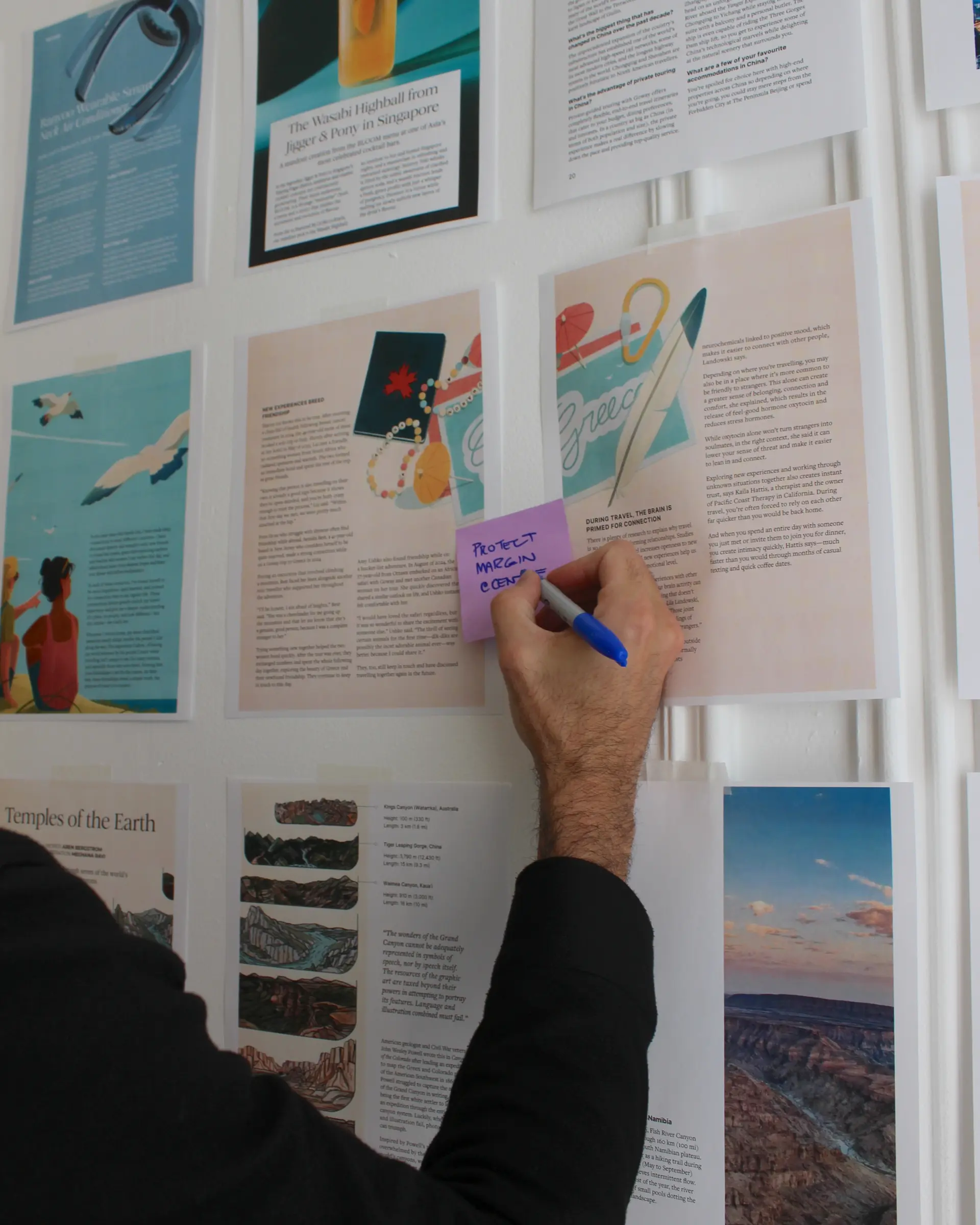 Hand writing a design note on a Post-it while revising an illustrated travel story layout on the Globetrotting pagination wall
