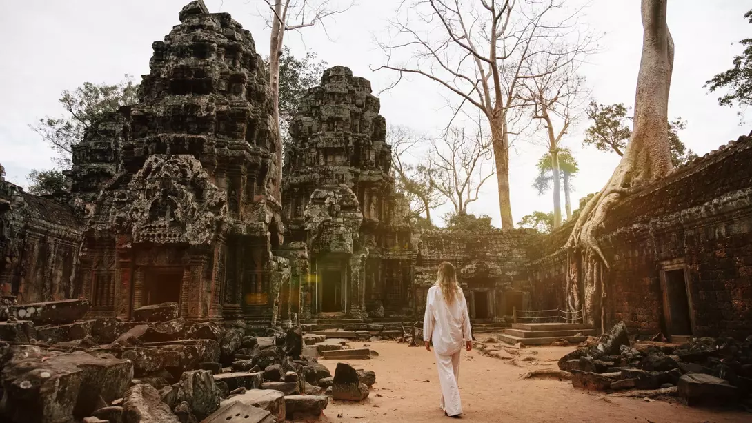 A person in white clothing walks through ancient temple ruins surrounded by trees and a sandy path.
