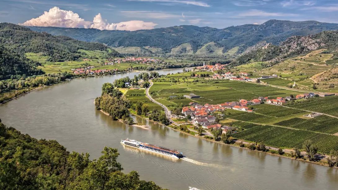 A scenic river winds through lush green hills and vineyards, with a boat navigating the water near a small village.