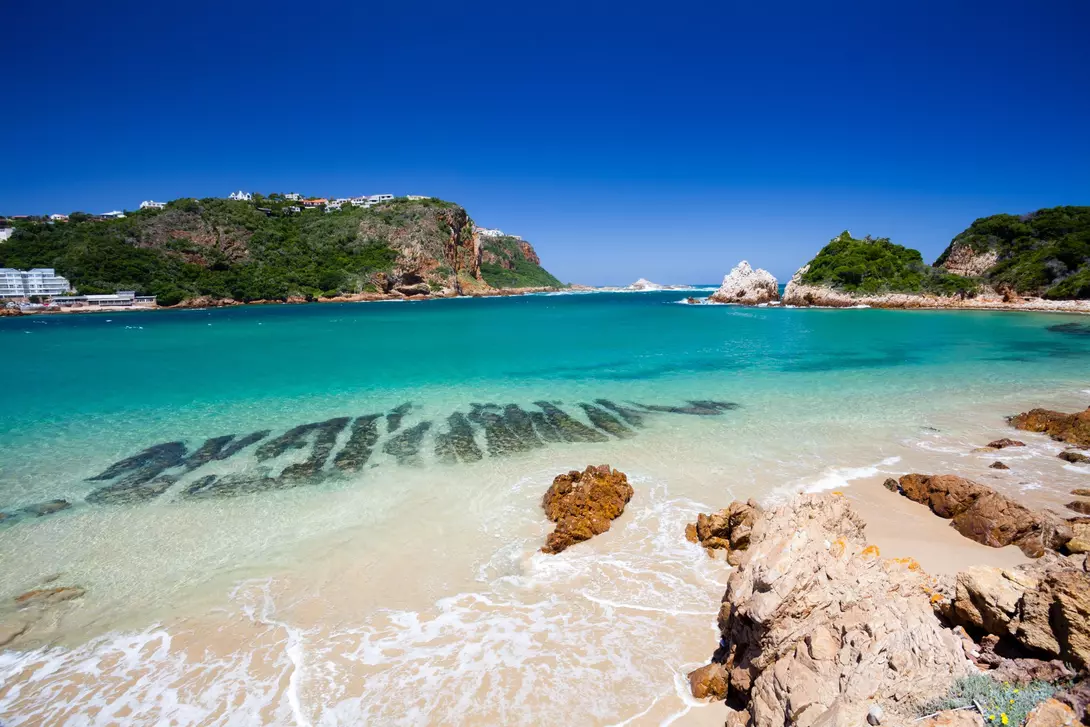 A beach in Knysna, Western Cape, South Africa. The beach is surrounded by lush green hills and has white sand and clear blue water.