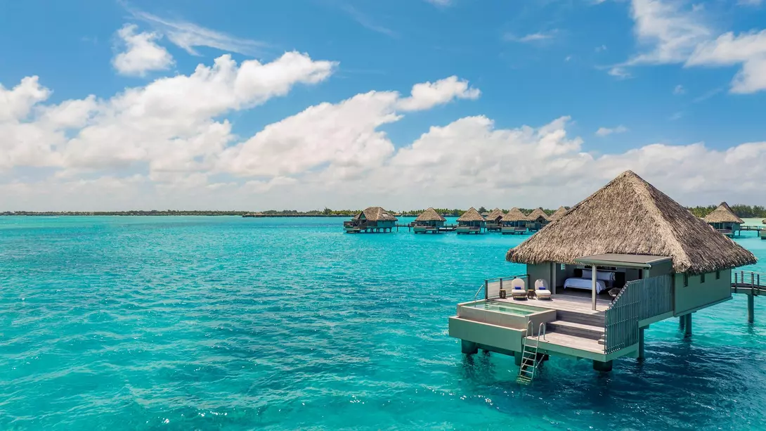 Bora Bora, French Polynesia Overwater bungalows with thatched roofs sit above bright turquoise water under a sunny sky, evoking a luxurious tropical island getaway.