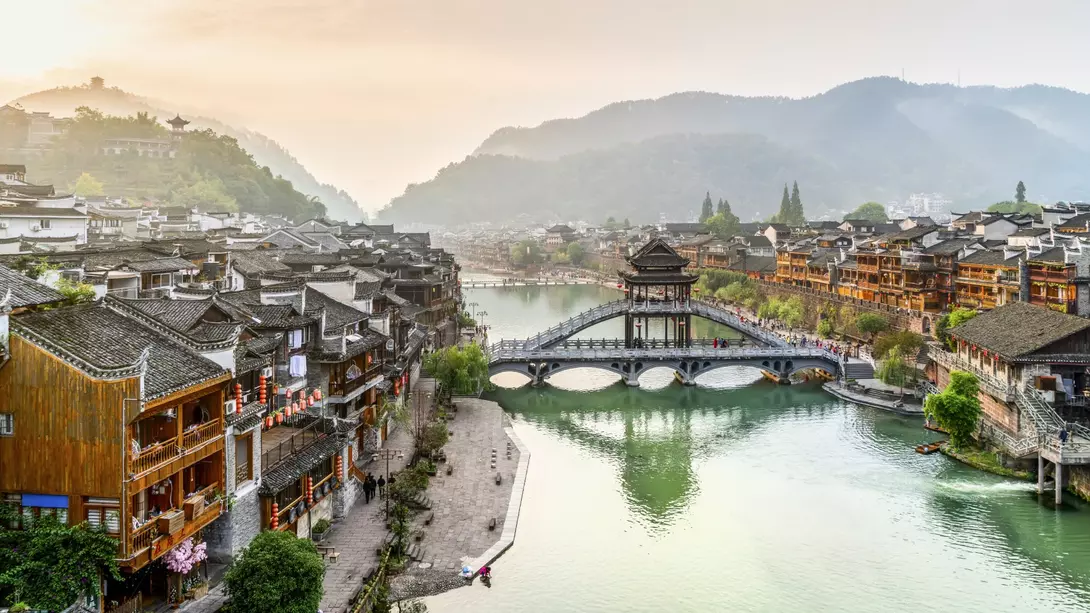 Scenic view of a river lined with traditional wooden buildings, a bridge, and mountains in the background at sunset.