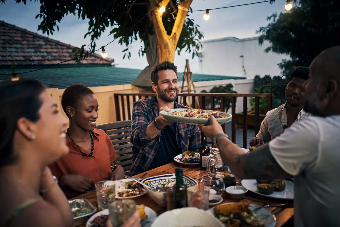 A group of friends enjoys dinner outdoors, sharing food and smiling at each other under string lights.