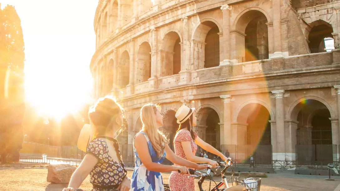 Three happy young women friends tourists with bikes at Colosseum in Rome, Italy at sunrise