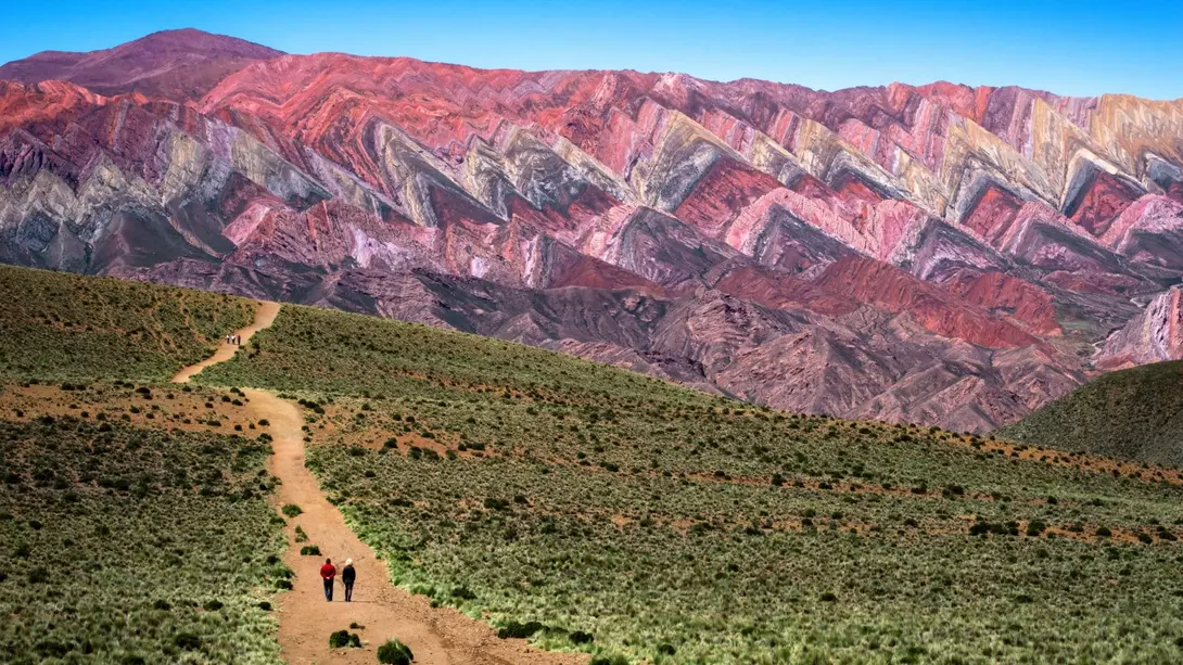 Two hikers walk along a dirt path with colorful striped mountains in the background under a clear blue sky.