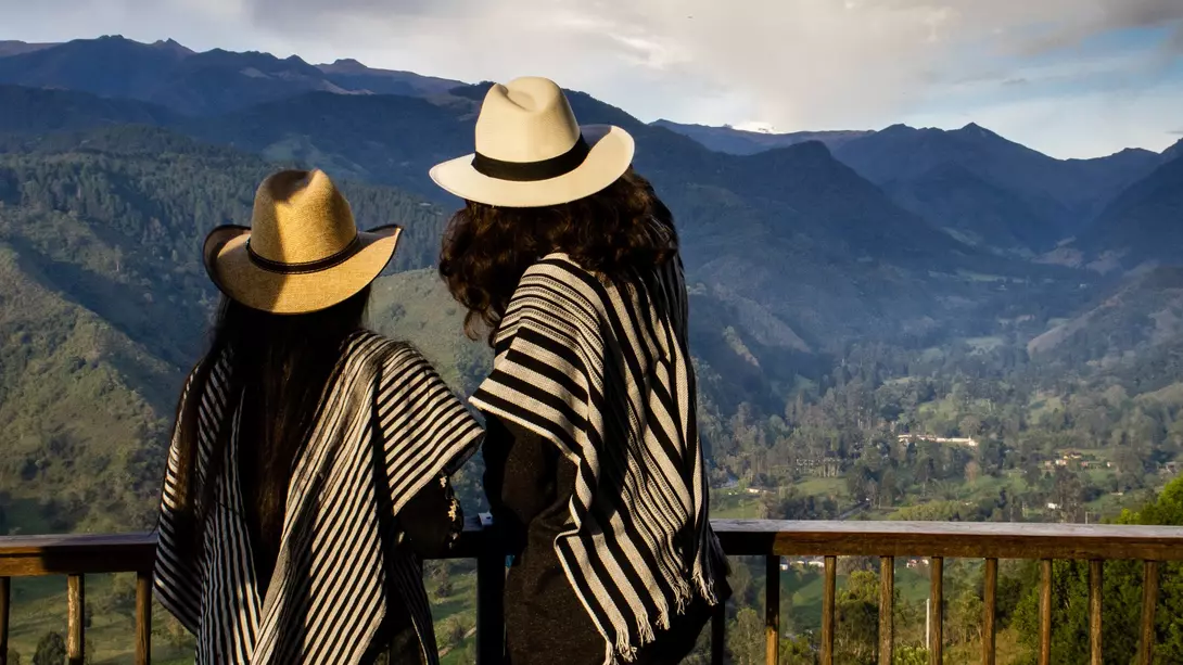 Two people in traditional ponchos and hats stand overlooking a mountainous landscape.