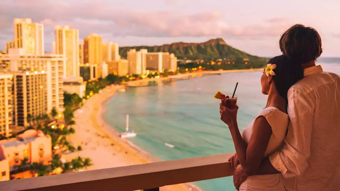 Embraced couple enjoying a sea-side view from a balcony, overlooking a city skyline and sunny sky.