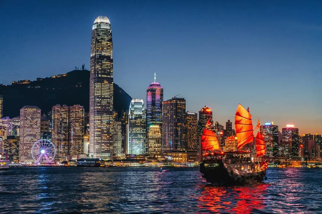 A junkboat on Hong Kong's Victoria Harbour at sunset against a backdrop of skyscrapers.