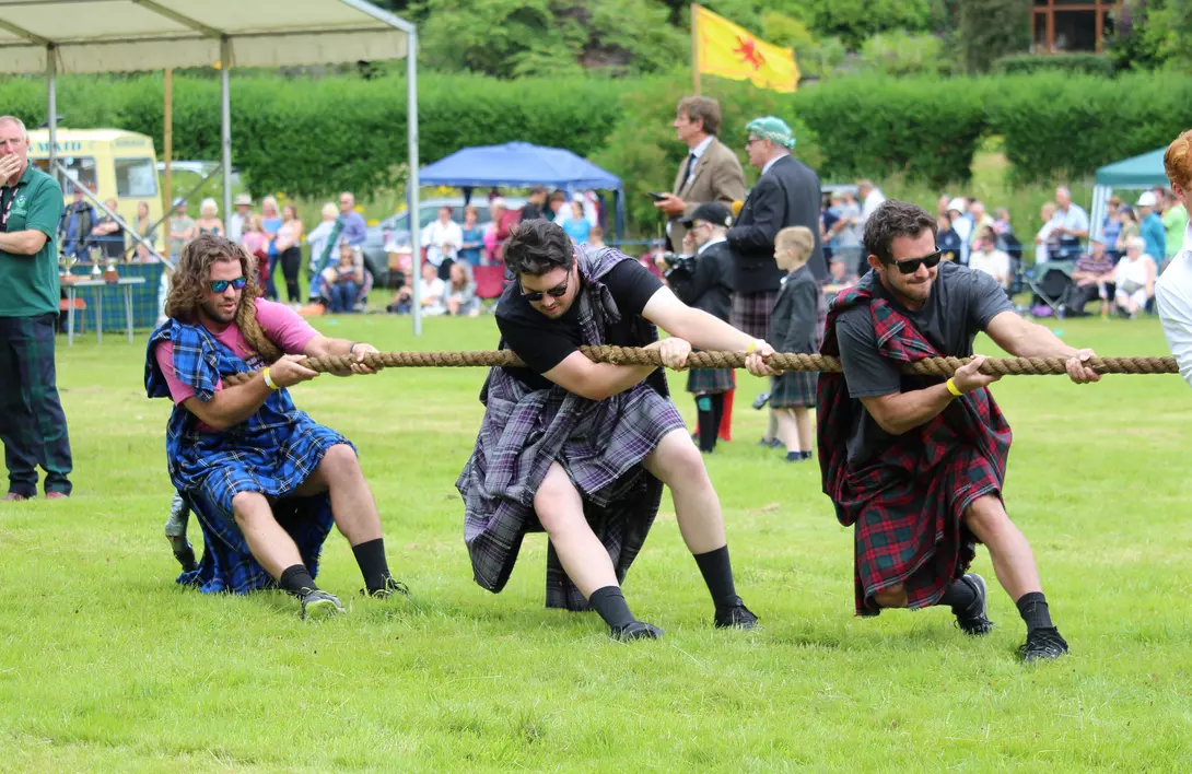 Three men in kilts are pulling a rope in a tug-of-war competition, with a crowd watching in the background.