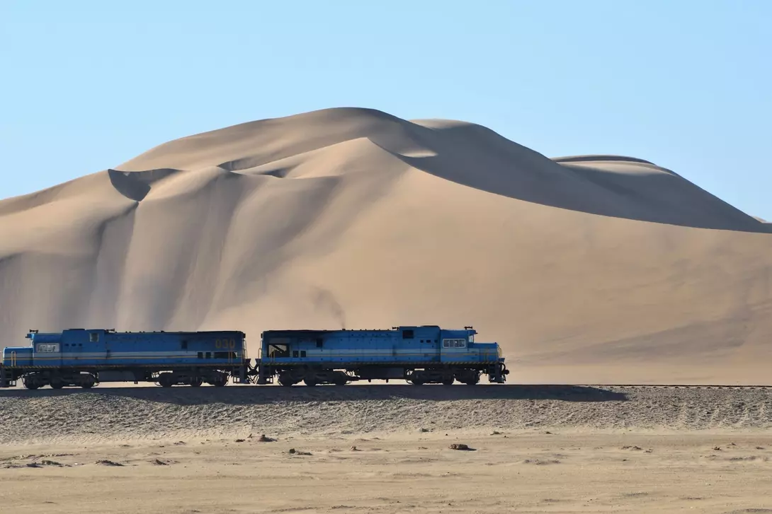 Two blue trains pass by a large sand dune under a clear blue sky.