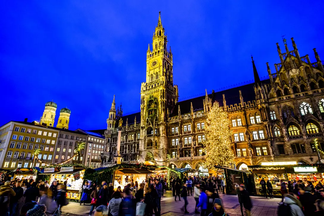 A bustling market scene at night with a tall clock tower and a lit Christmas tree in the background.