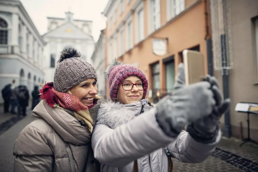 Two women in winter attire taking a selfie on a city street with historic buildings in the background.