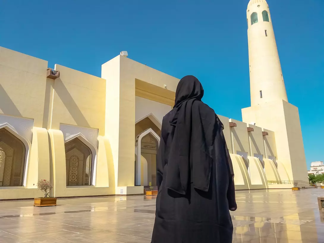 A person in a black abaya stands before a large mosque with a tall minaret under a blue sky.