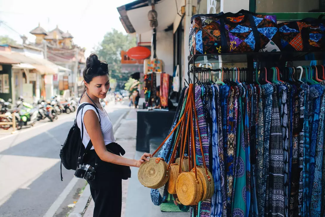 A woman examines woven bags in a market, with colorful fabrics hanging nearby and a street scene in the background.