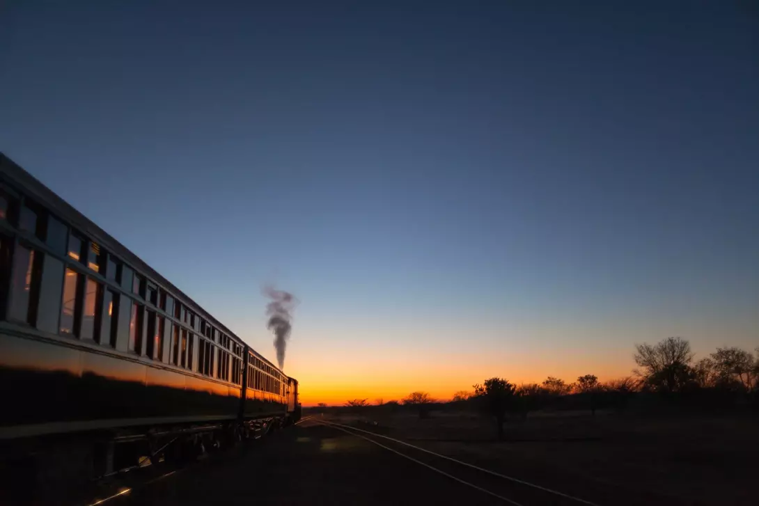 A steam train travels along tracks at sunset, with vibrant colors in the sky and a silhouette of trees in the distance.