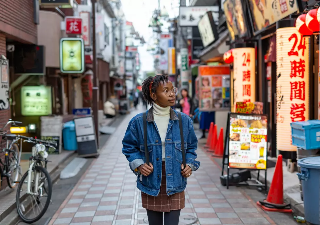 A person in a denim jacket stands in a vibrant urban alley filled with shops and neon signs.