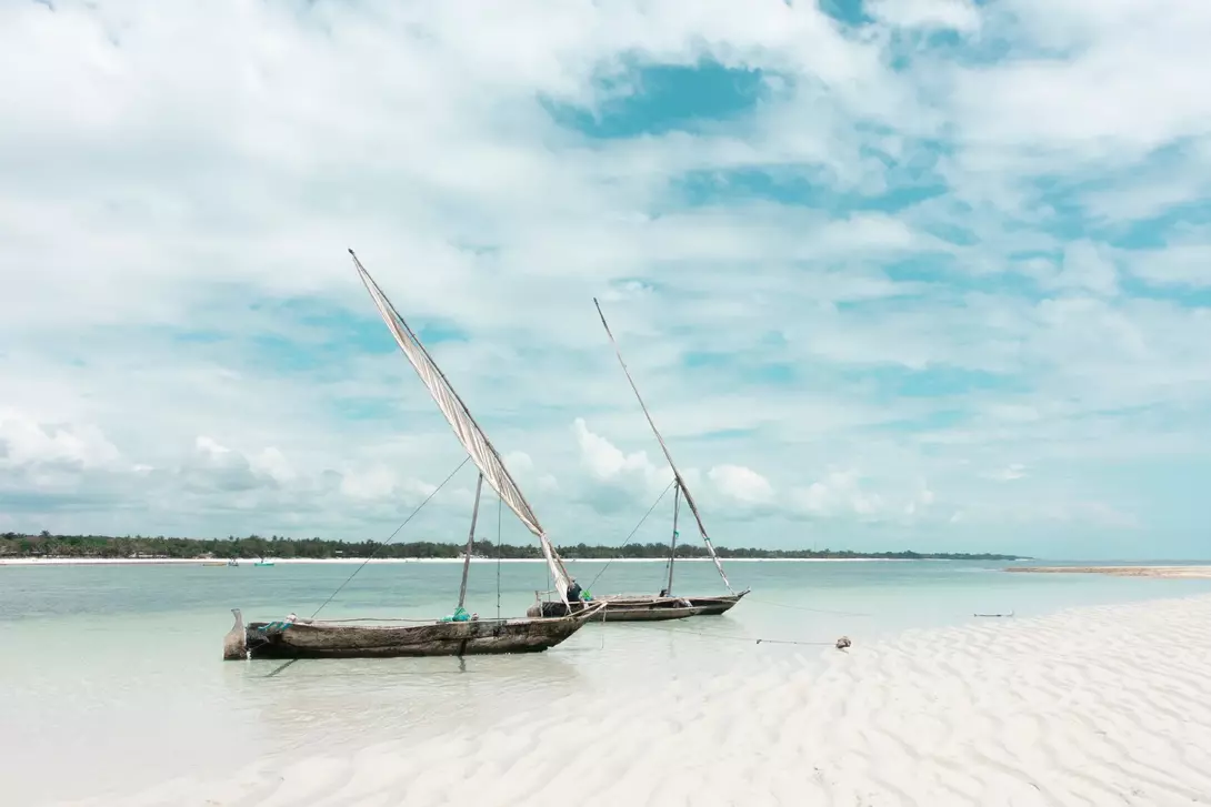 Two traditional sailboats on a calm beach, with soft sand and a cloudy sky in the background.
