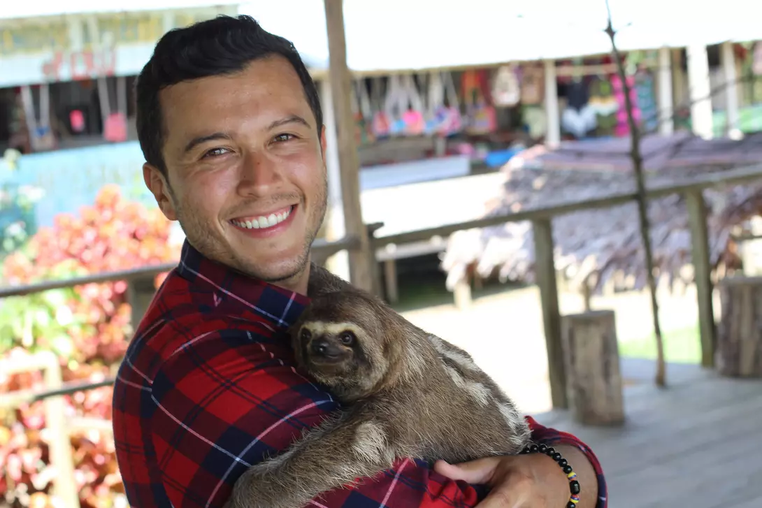 Smiling person in a plaid shirt holds a sloth in their arms, with a colorful background of a shop and plants.