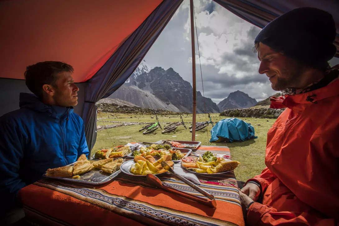 Two people sit inside a tent with a table of food, overlooking a scenic mountain landscape.