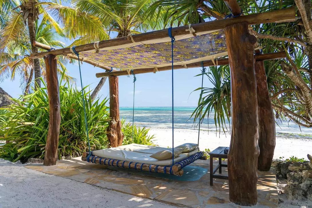 A shaded hammock swing under palm trees on a beach, with clear water and white sand in the background.