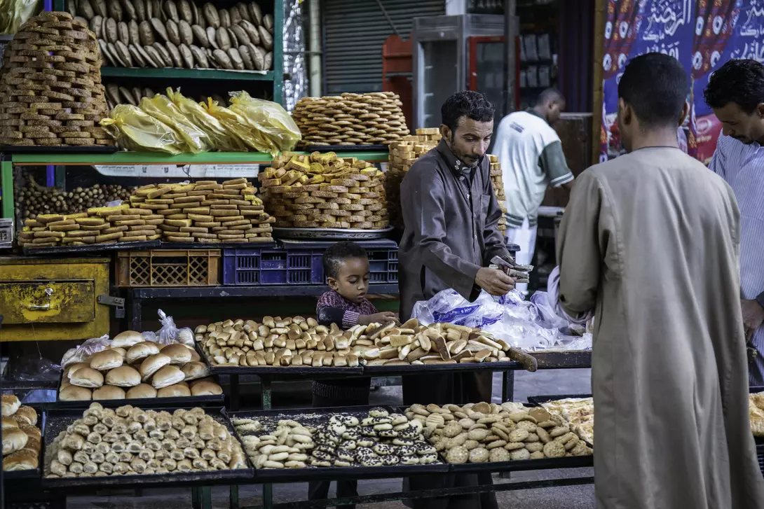 A busy market stall displays various breads and pastries, with a child watching and adults selecting items.