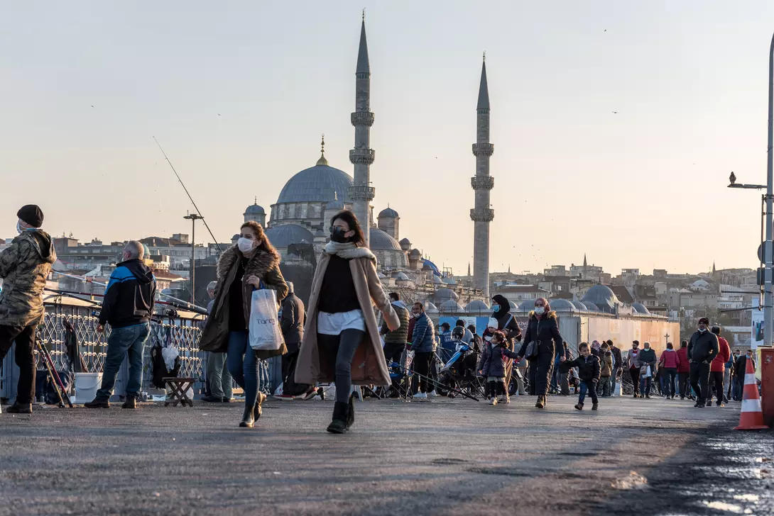 Crowds of people walk along a busy bridge with the skyline and mosque minarets in the background at sunset.