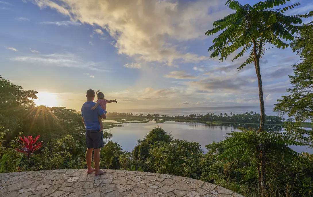 A parent holds a child while gazing at a scenic view of a lake and lush landscape under a bright sky.