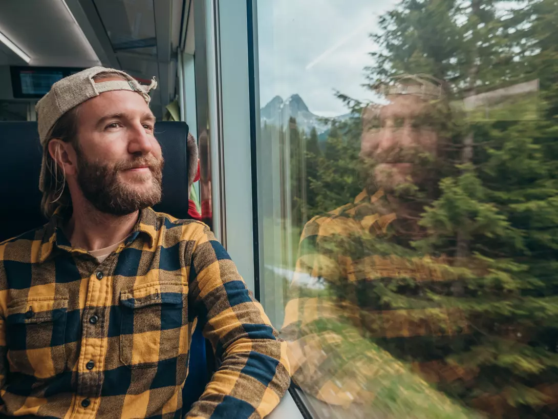 A man in a plaid shirt and cap gazes out the train window, with mountains and trees reflected in the glass.