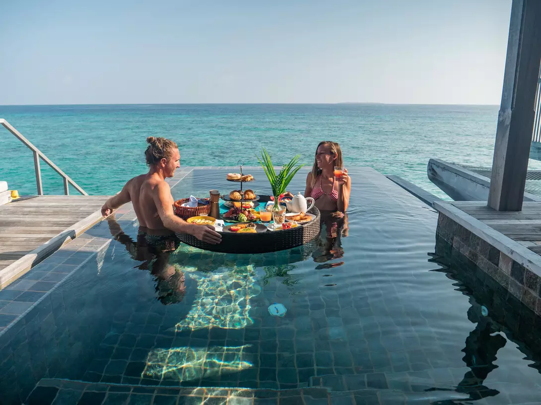 A couple relaxes in a pool, enjoying a floating breakfast with scenic ocean views.