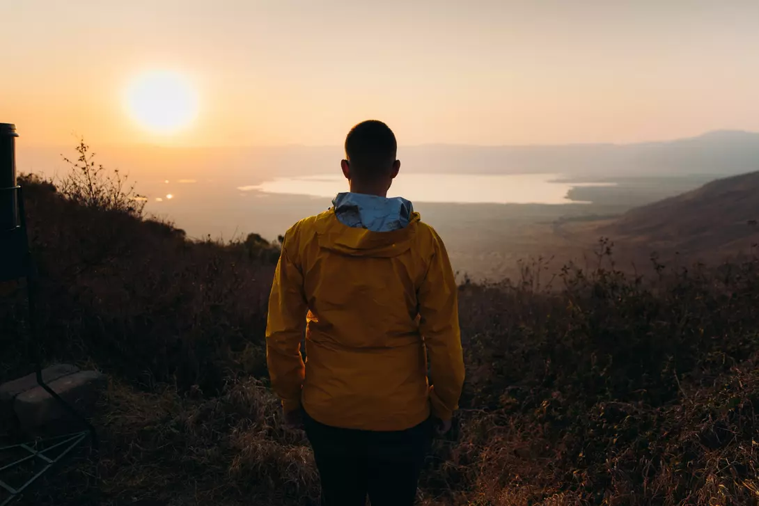 A person in a yellow jacket stands facing a sunset over a lake, surrounded by hills and vegetation.