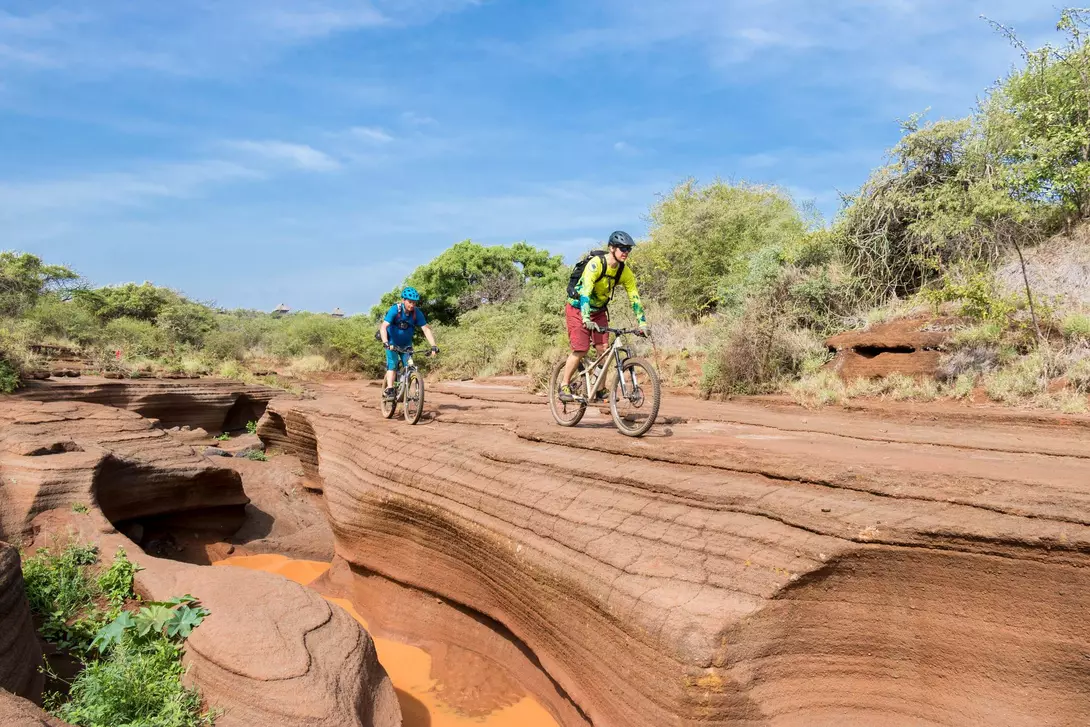 Two mountain bikers ride along a rocky, terraced landscape under a blue sky, surrounded by green vegetation.
