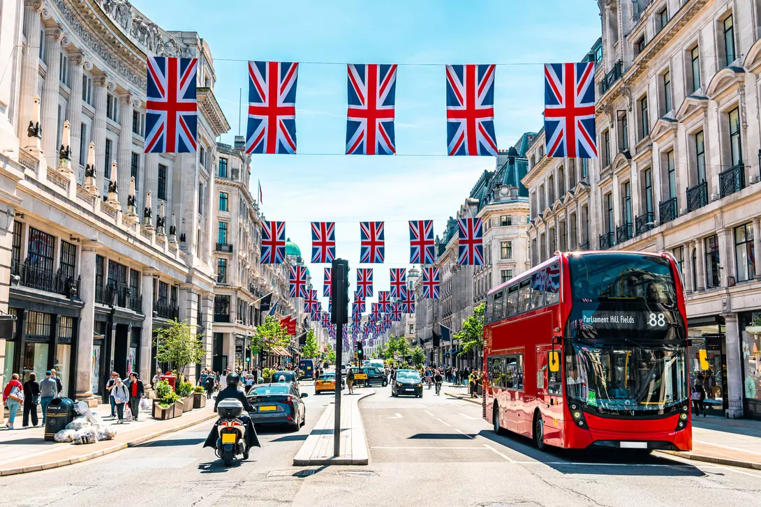 A busy street lined with shops, featuring red double-decker buses and decorative Union Jack flags overhead.