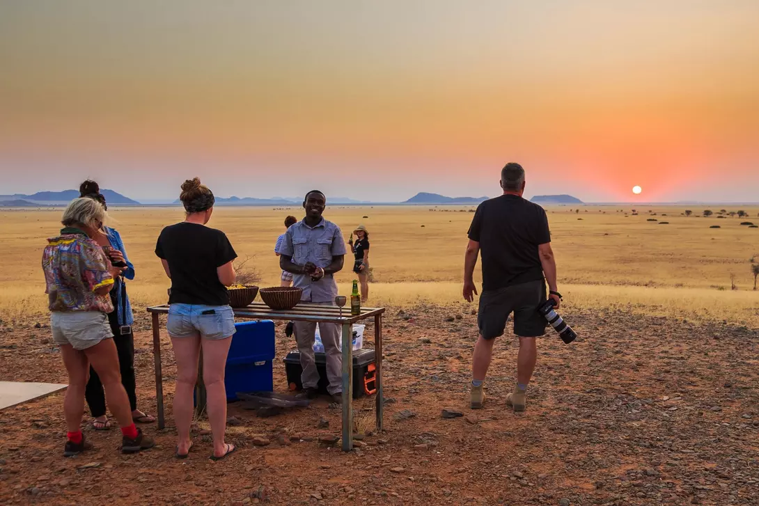 People gather around a table in a sunset-lit desert, enjoying a meal while a photographer captures the moment.
