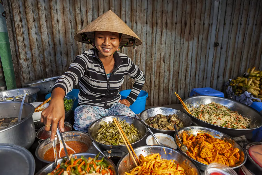 A smiling woman in a conical hat serves dishes from a variety of bowls at a vibrant market stall.