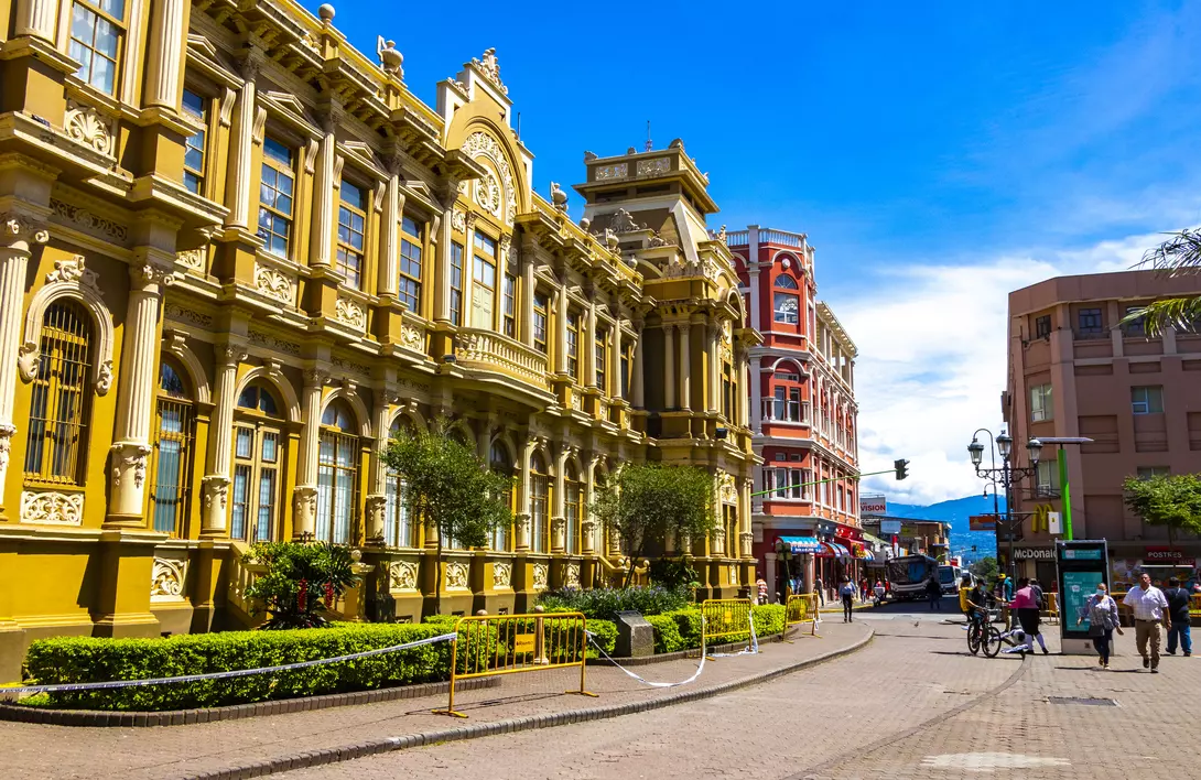 Historic buildings line a sunny street, with greenery and pedestrians enjoying the vibrant urban scene.
