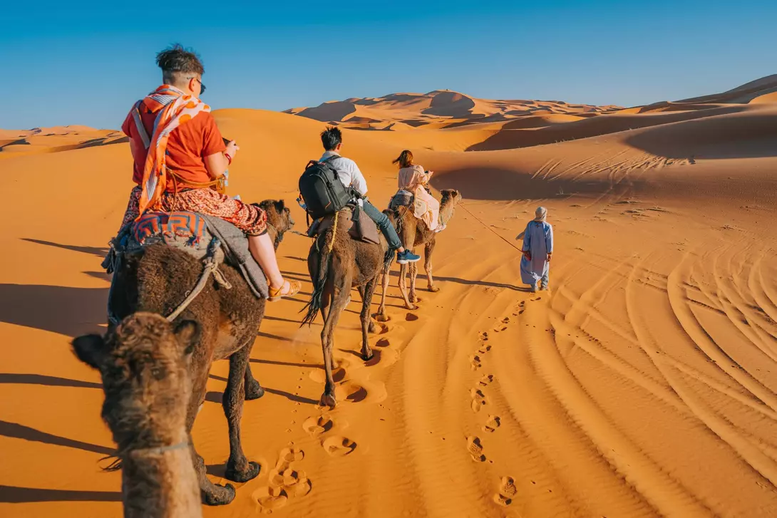 A group of people riding camels through golden sand dunes under a clear blue sky.