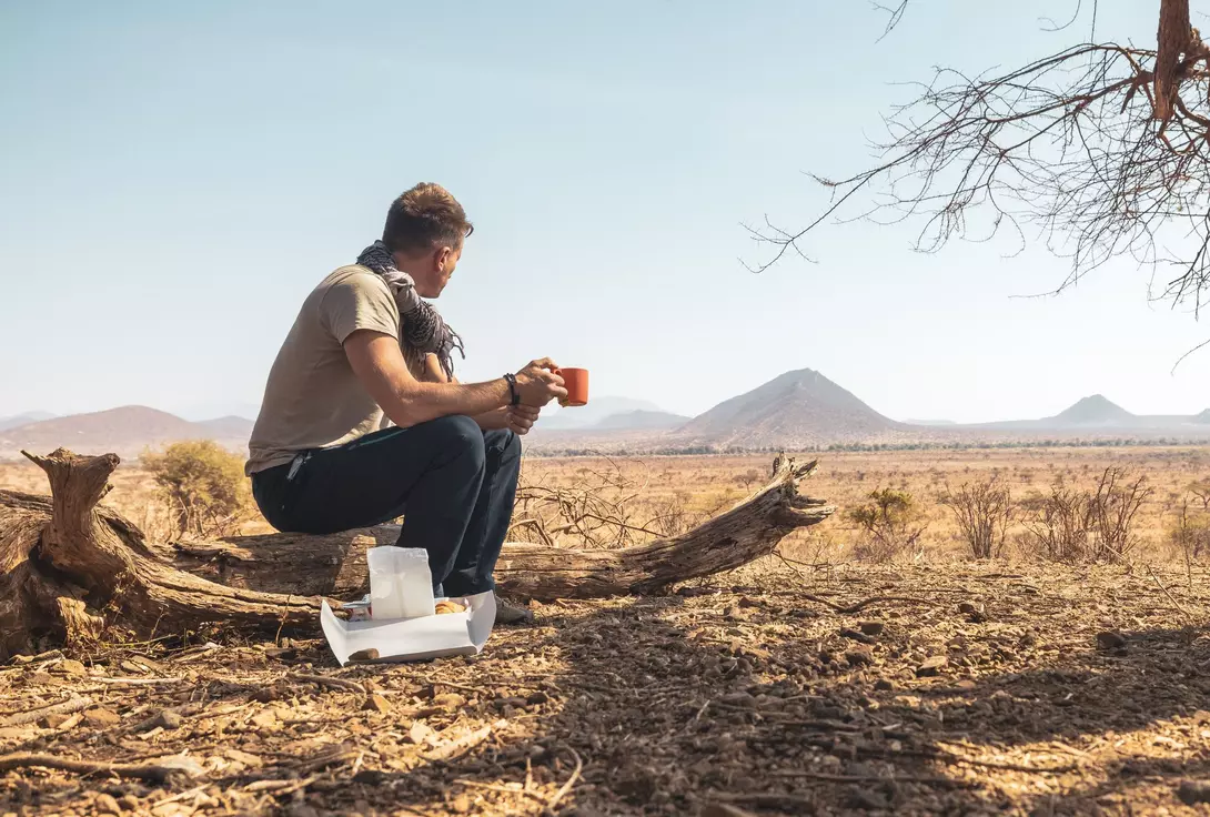 A person sits on a log in a vast, arid landscape, holding a cup and looking out at distant mountains.