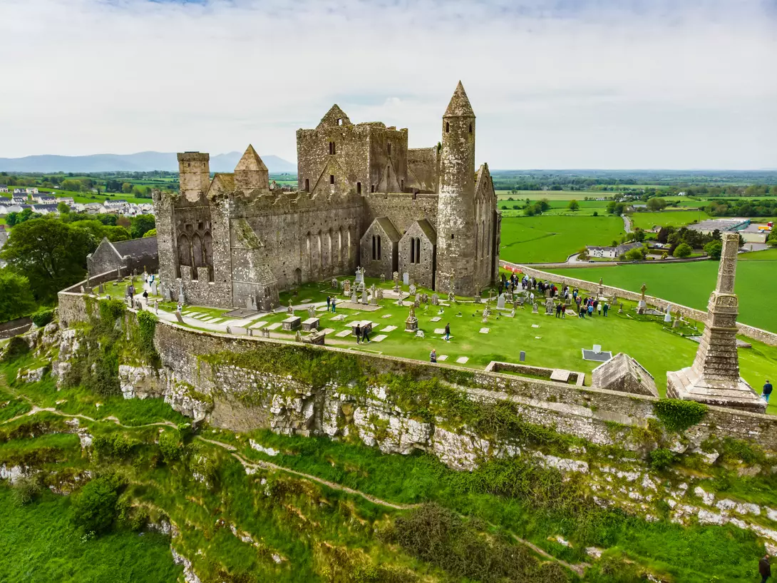 Aerial view of a historic castle surrounded by green fields and gravestones on a hilltop.