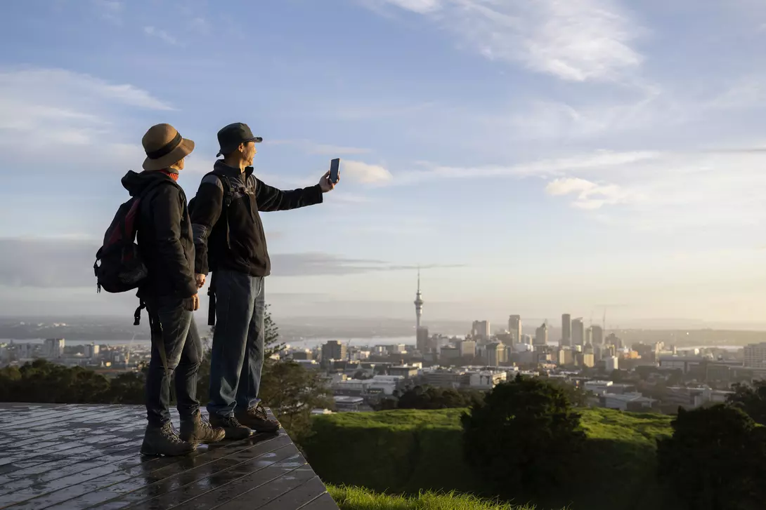 Two people take a selfie with a city skyline in the background during sunset, standing on a green hill.