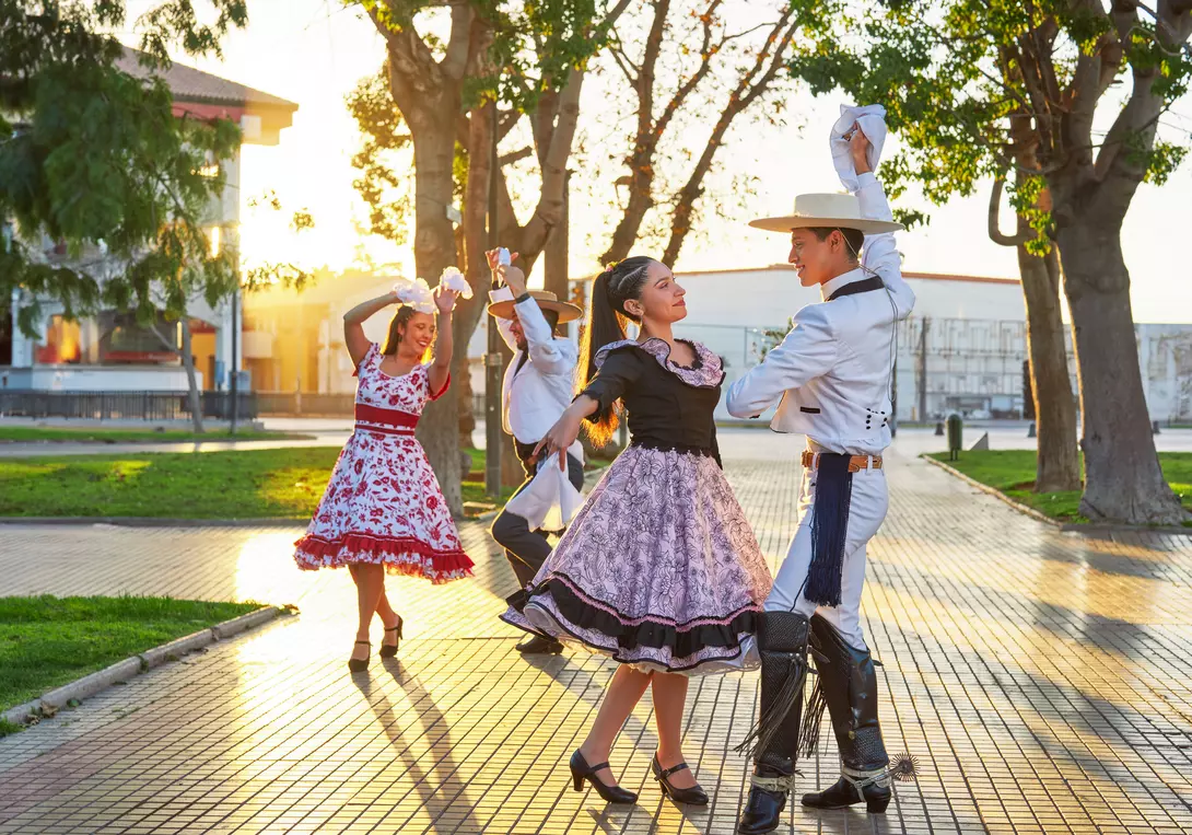 A couple dances in traditional attire outdoors, with more dancers in the background during sunset.